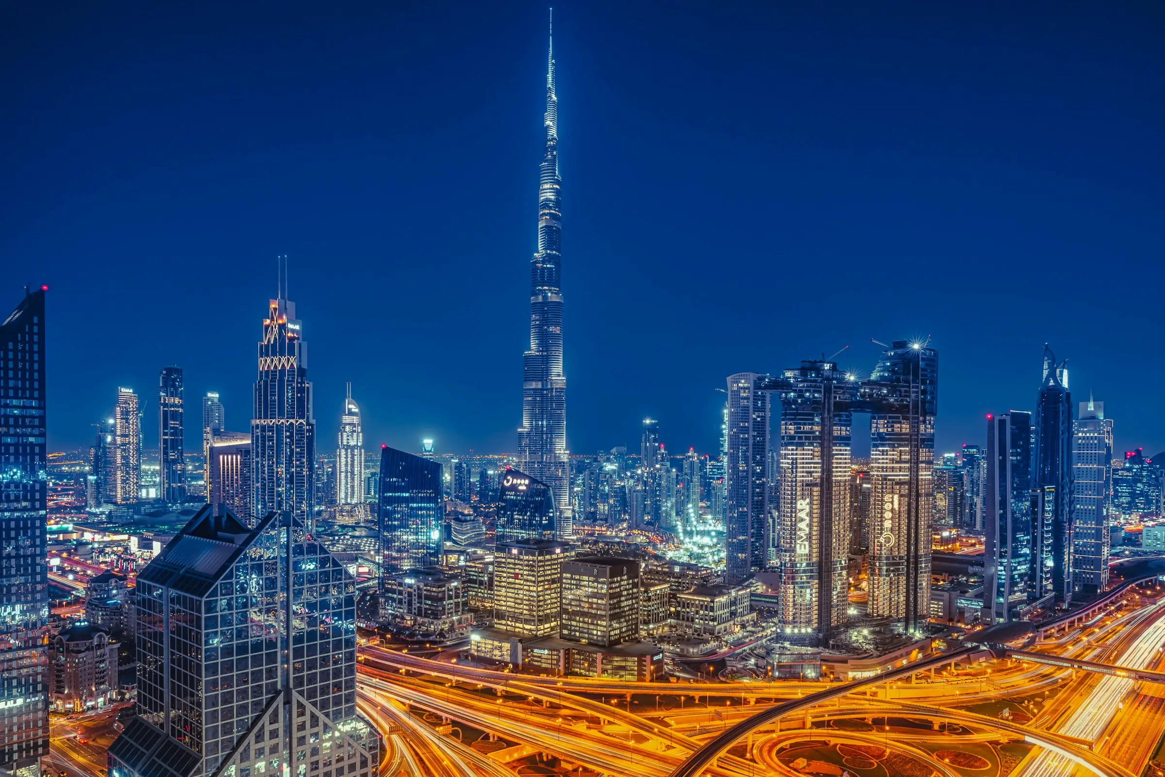 Downtown Dubai at blue hour, the Burj Khalifa lit above the towers and the Sheikh Zayed Road interchange traced in light trails