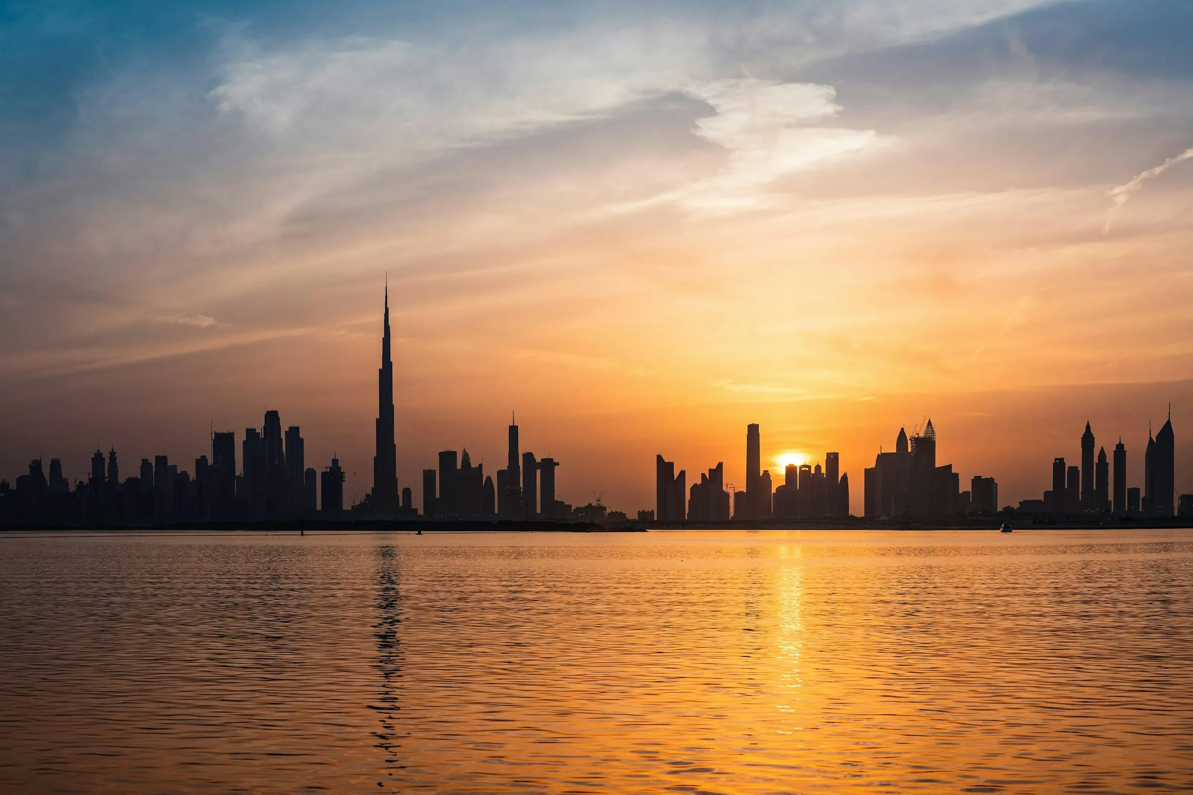 The Dubai skyline in silhouette at sunset, with the Burj Khalifa rising above the towers and the sun reflected across the water